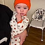 Malcolm is registered to the contest to win money with this photo: baby, child, infant, halloween, pumpkin_hat, onesie, hand, chewing, adult_hand, high_chair, kitchen, cabinet, floor, person, indoor, cute, costume, clothing, face, portrait