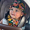 baby, child, stroller, hat, bucket_hat, floral_pattern, colorful_clothing, seatbelt, infant, cute, portrait, smile, face, earrings, closeup, outdoor, sunlight, comfort, baby_seat, expression