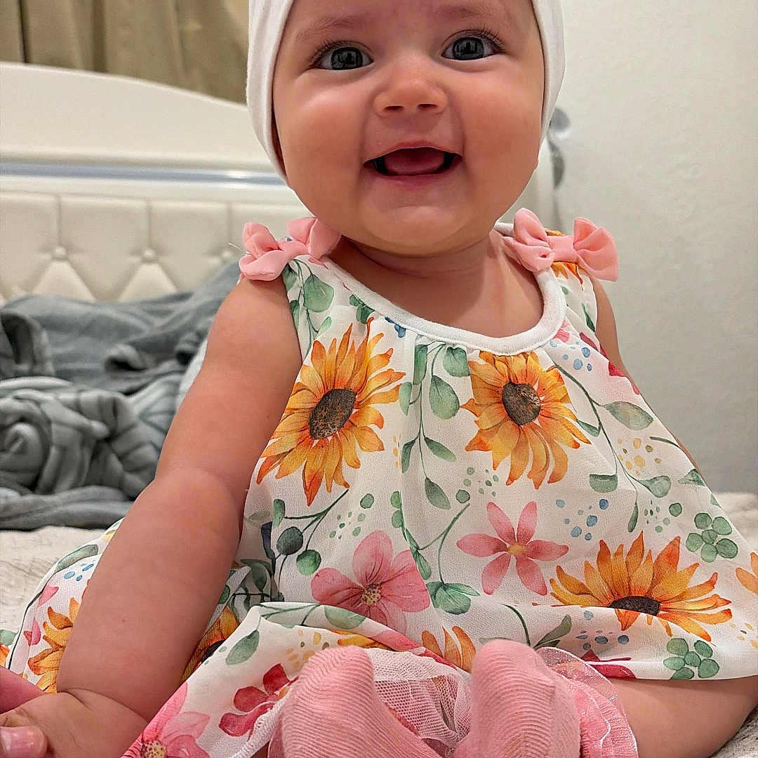 Shaddaimarie is registered to the contest to win money with this photo: baby, child, smiling, sitting, dress, headband, flower_pattern, sunflower, pink_socks, blanket, bedroom, curtain, happy, infant, indoor, cute, portrait, person, toddler, clothing