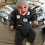 baby, child, smiling, happy, sweater, hat, pom_poms, sheep_pattern, couch, blanket, indoor, cozy, clothing, footwear, face, person, portrait, seated, wall, home