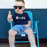 Emylio a rejoint le concours — aidez-le/la à gagner de superbes lots ! toddler, child, sunglasses, blue_chair, shorts, tshirt, white_sneakers, pointing, indoor, casual_clothing, confident, pose, person, footwear, short_hair, seat, young_child, fashion, cool, portrait