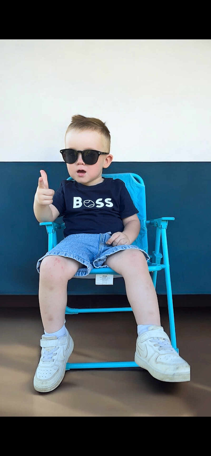 Emylio a rejoint le concours — aidez-le/la à gagner de superbes lots ! toddler, child, sunglasses, blue_chair, shorts, tshirt, white_sneakers, pointing, indoor, casual_clothing, confident, pose, person, footwear, short_hair, seat, young_child, fashion, cool, portrait