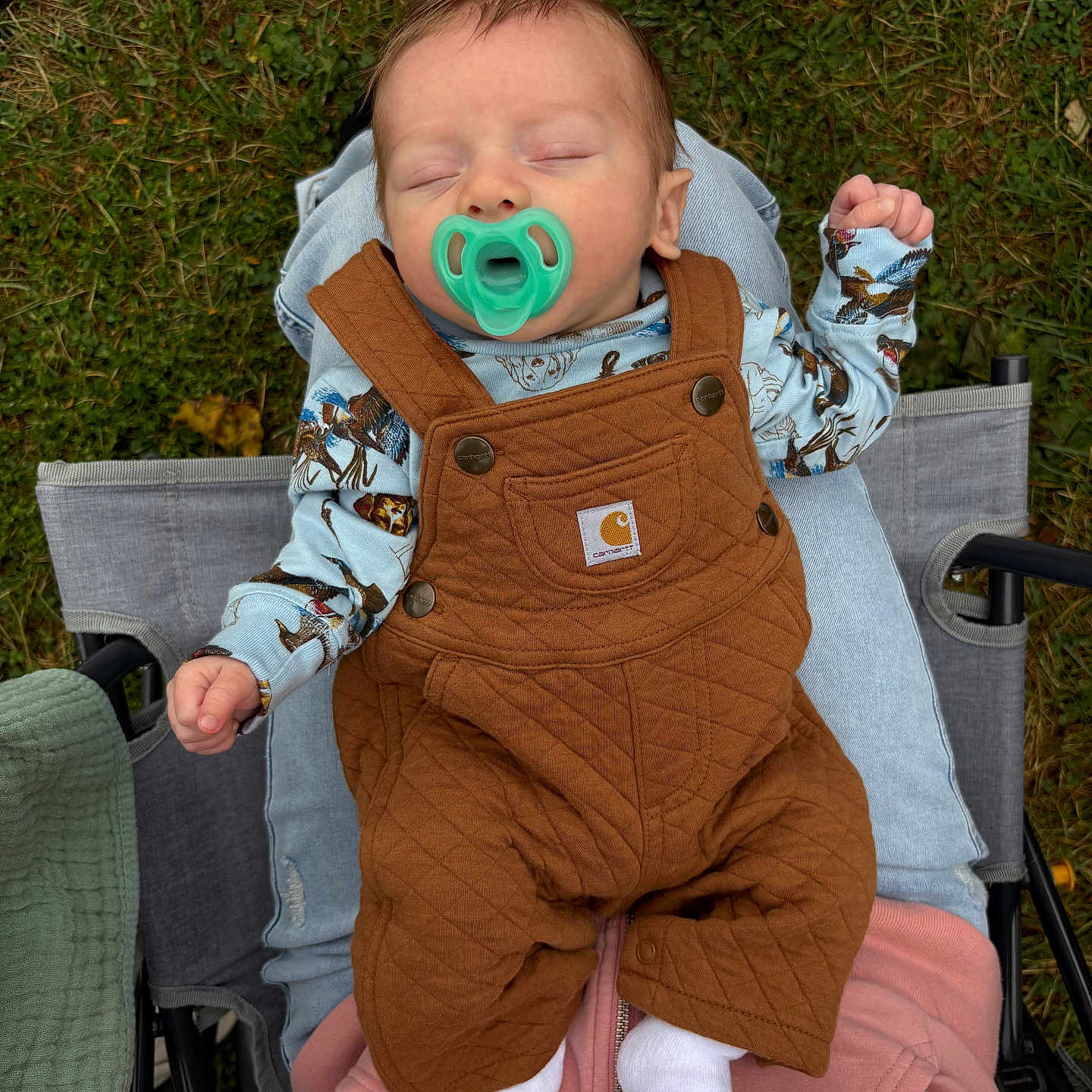 Kane is registered to the contest to win money with this photo: baby, blue_shirt, brown_clothing, casual, comfort, footwear, grass, hand, infant, lap, nature, outdoor, overalls, pacifier, person, quilted, relaxation, sleeping, socks, stroller