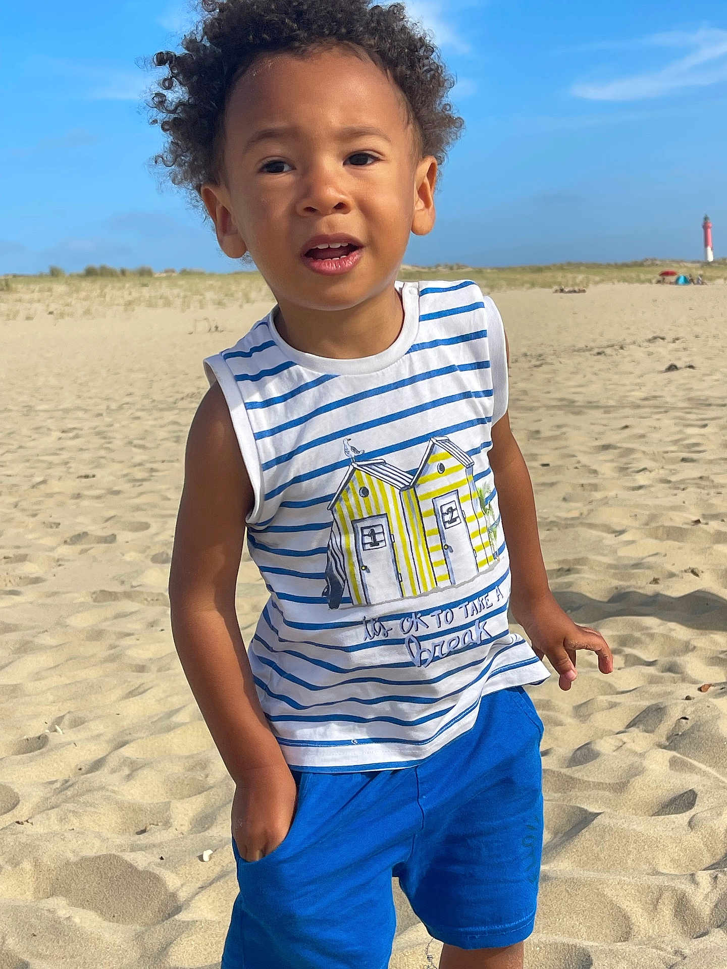 Marlon participe au concours pour gagner de l'argent avec cette photo : child, beach, sand, blue_sky, striped_shirt, blue_shorts, curly_hair, casual_pose, summer_clothing, outdoor, sunny, young_boy, standing, portrait, daytime, nature, vacation, sand_dunes, happy, exploring