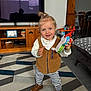 toddler, child, toy, phone, living_room, rug, patterned_rug, brown_shoes, striped_pants, brown_vest, white_shirt, smiling, happy, indoor, furniture, television, wooden_cabinet, curtains, home_decor, person
