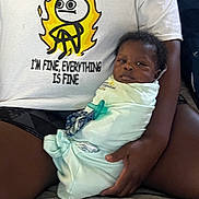 Eli is registered to the contest to win money with this photo: baby, swaddled, blanket, pacifier, adult, tshirt, cartoon, text, indoor, person, holding, seated, legs, hand, expression, face, clothing, casual, comfort, candid