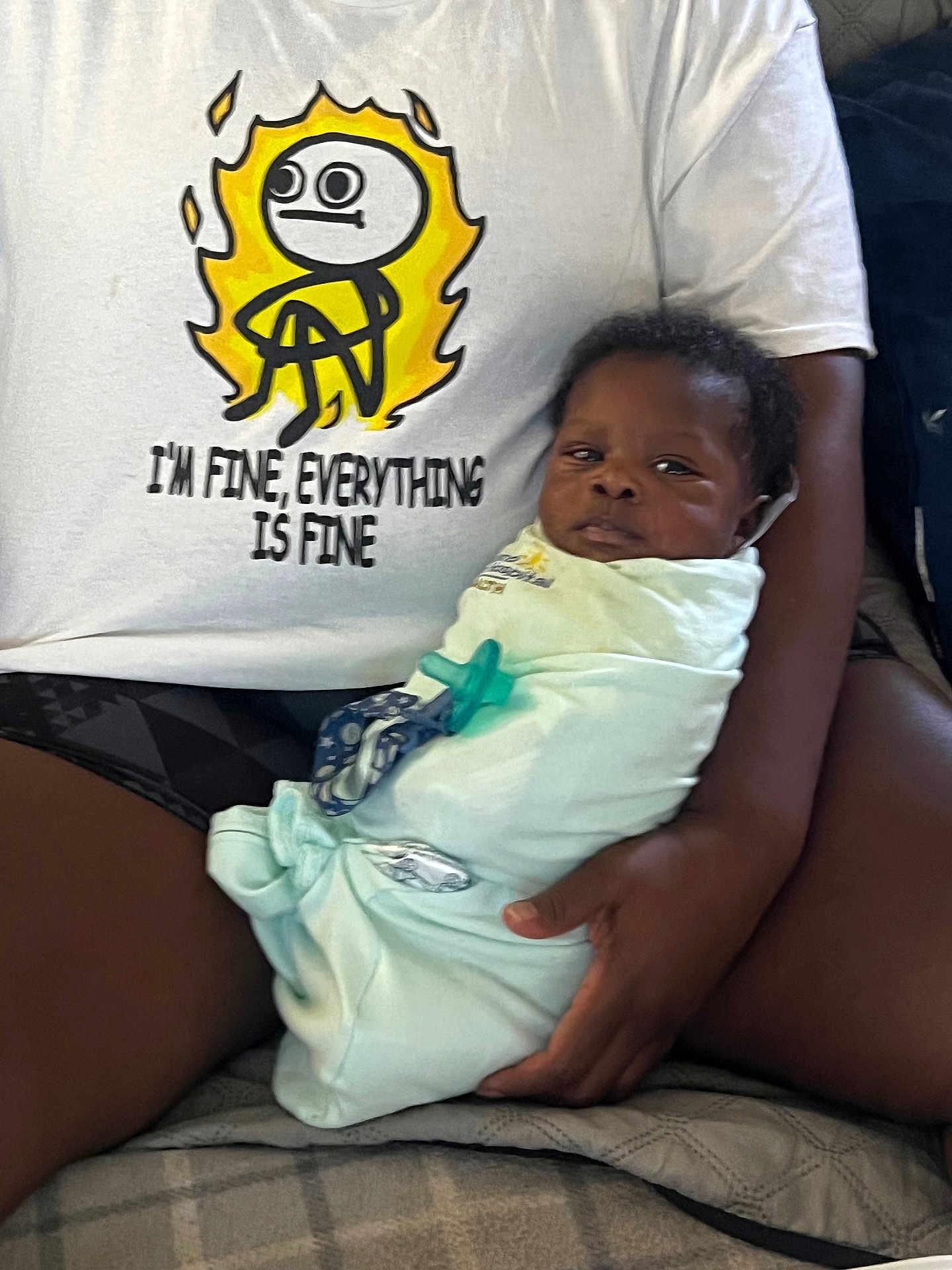 Eli is registered to the contest to win money with this photo: baby, swaddled, blanket, pacifier, adult, tshirt, cartoon, text, indoor, person, holding, seated, legs, hand, expression, face, clothing, casual, comfort, candid