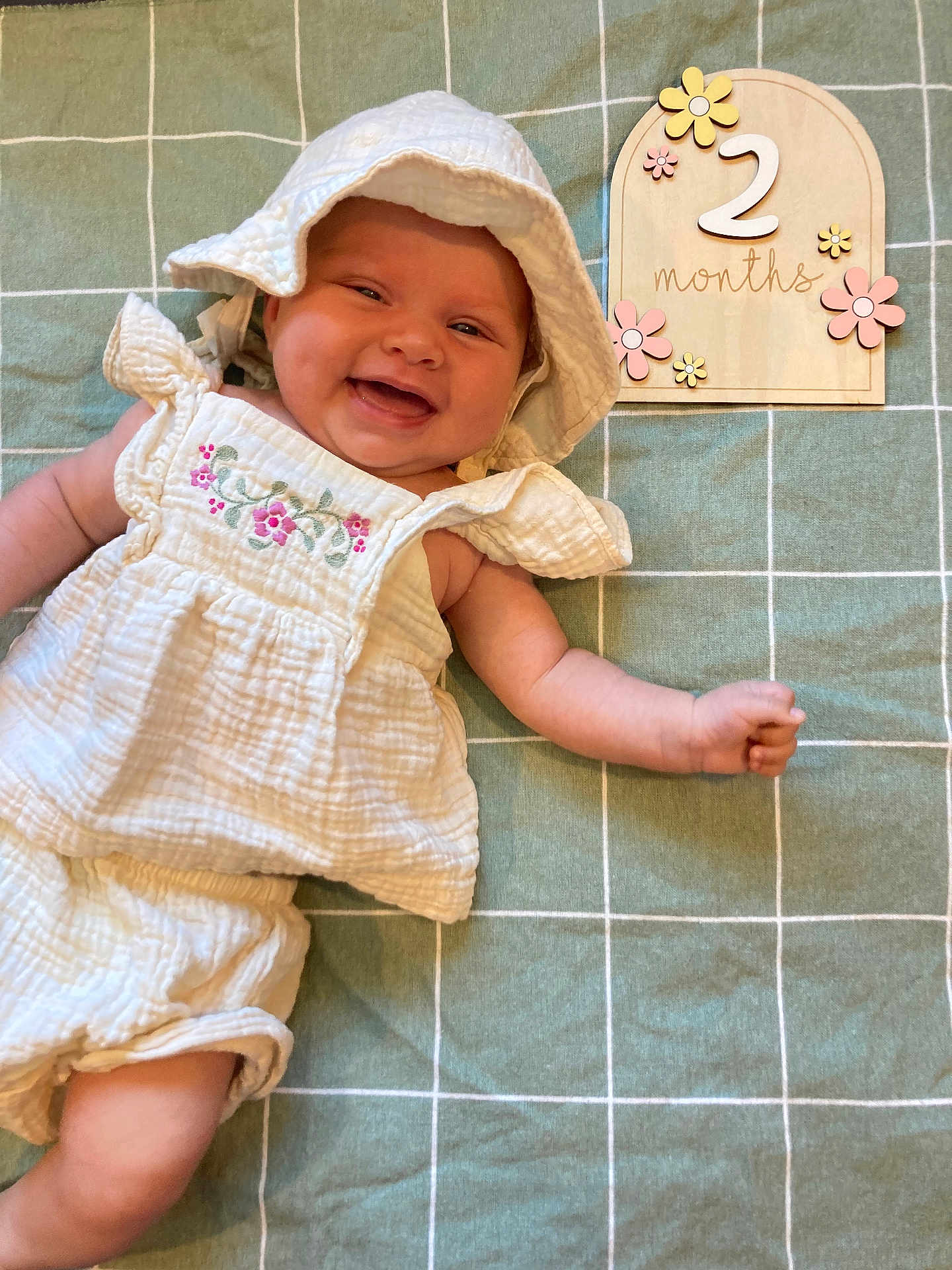 Sage is registered to the contest to win money with this photo: baby, infant, smiling, white_clothing, hat, blanket, green, checkered, wooden_sign, flowers, months, cute, happy, lying_down, arm, skin, child, portrait, closeup, adorable