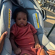 Micah is registered to the contest to win money with this photo: baby, infant, car_seat, child, outdoor, sunlight, seat, clothing, person, hand, texture, warning_label, urban, sidewalk, curb, street, window, shadow, brown_skin, expression