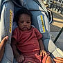 baby, infant, car_seat, child, outdoor, sunlight, seat, clothing, person, hand, texture, warning_label, urban, sidewalk, curb, street, window, shadow, brown_skin, expression