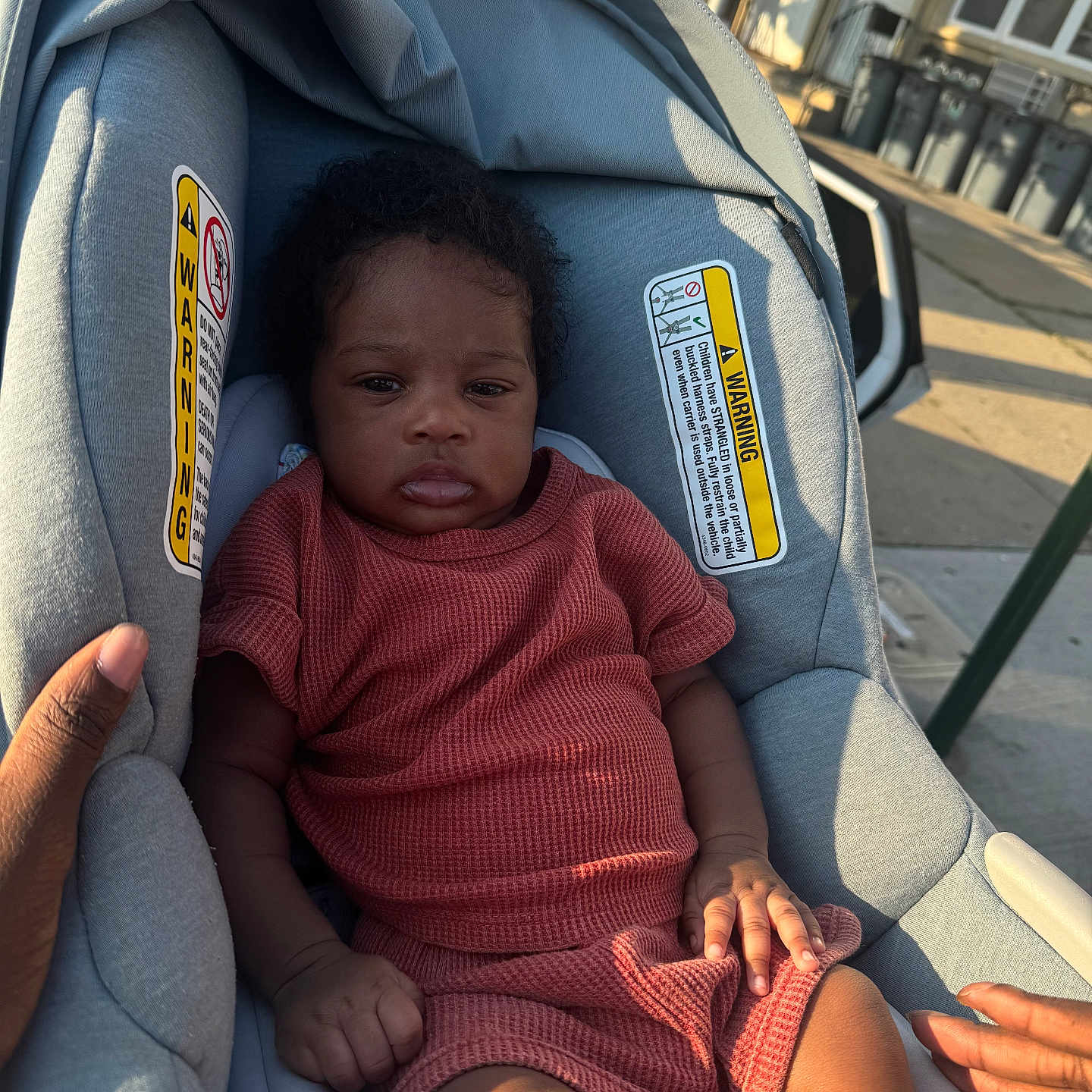 Micah is registered to the contest to win money with this photo: baby, brown_skin, car_seat, child, clothing, curb, expression, hand, infant, outdoor, person, seat, shadow, sidewalk, street, sunlight, texture, urban, warning_label, window