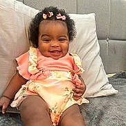 Gabriella is registered to the contest to win money with this photo: baby, smiling, cute, child, infant, sitting, bed, pillow, hair_bow, floral_outfit, happy, indoors, skin, legs, white_socks, curly_hair, couch, comfort, portrait, relaxing