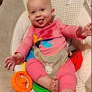 Madelyn is registered to the contest to win money with this photo: baby, infant, smiling, tongue_out, pink_clothing, baby_seat, toys, colorful_toys, happy, cute, child, indoors, playful, cushion, feet, hands, person, face, head, sitting