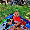 baby, child, person, outdoor, grass, blanket, toy, bottle, scooter, group, wheelchair, balloons, casual_clothing, park, sunlight, people, seated, greenery, shadows, gathering