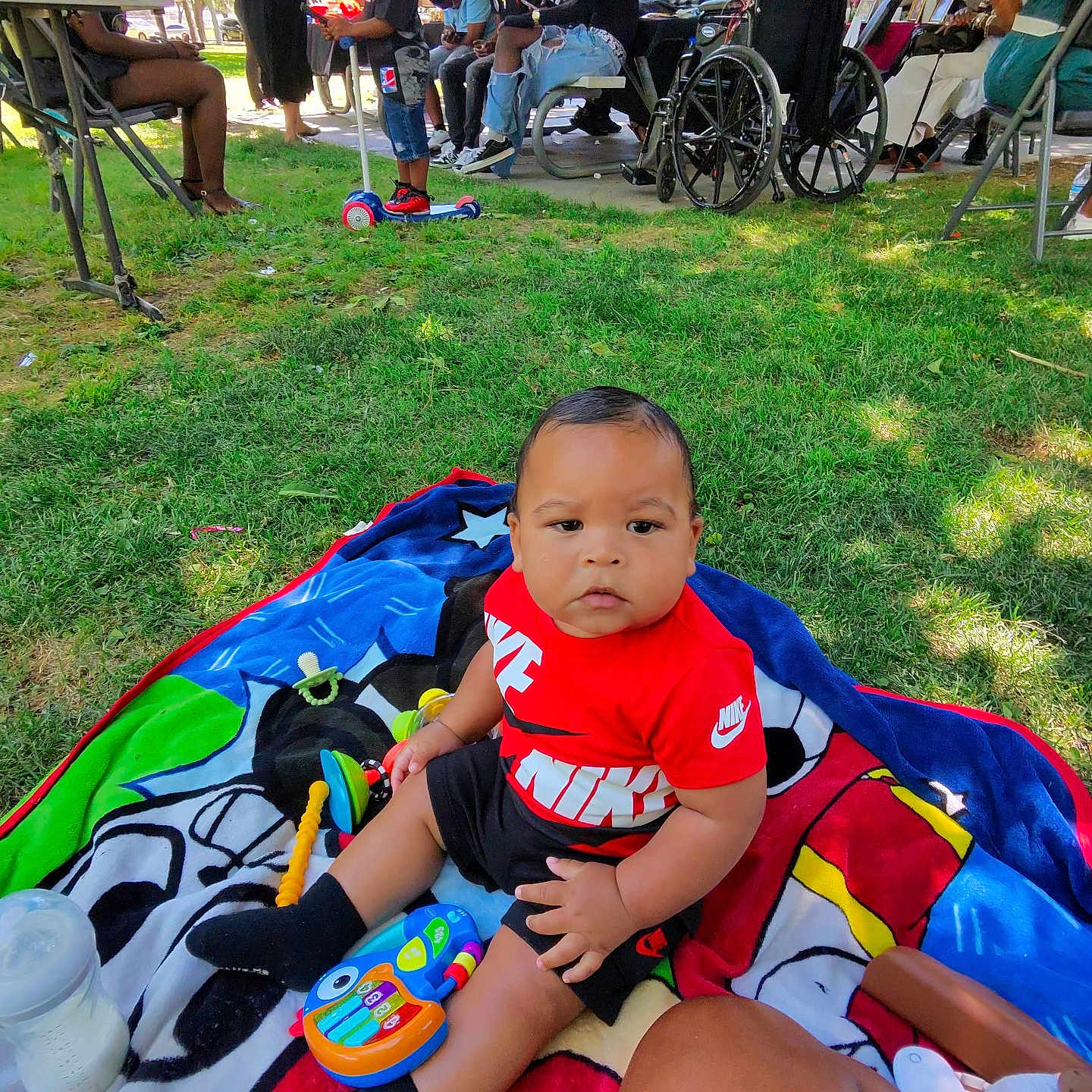 Behr is registered to the contest to win money with this photo: baby, child, person, outdoor, grass, blanket, toy, bottle, scooter, group, wheelchair, balloons, casual_clothing, park, sunlight, people, seated, greenery, shadows, gathering