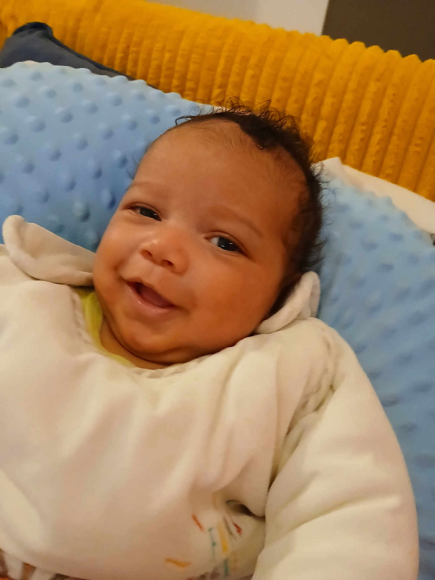 Martin a rejoint le concours — aidez-le/la à gagner de superbes lots ! baby, smiling, infant, face, white_clothing, pillow, blue_pillow, yellow_cushion, cozy, indoors, cute, child, portrait, resting, comfortable, soft_texture, young_child, happy, closeup, warm_colors