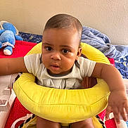 Ean is registered to the contest to win money with this photo: baby, child, infant, yellow_cushion, seat, arm, face, curious, indoor, bed, blanket, stuffed_toy, blue_toy, clothing, person, skin, head, expression, cute, home