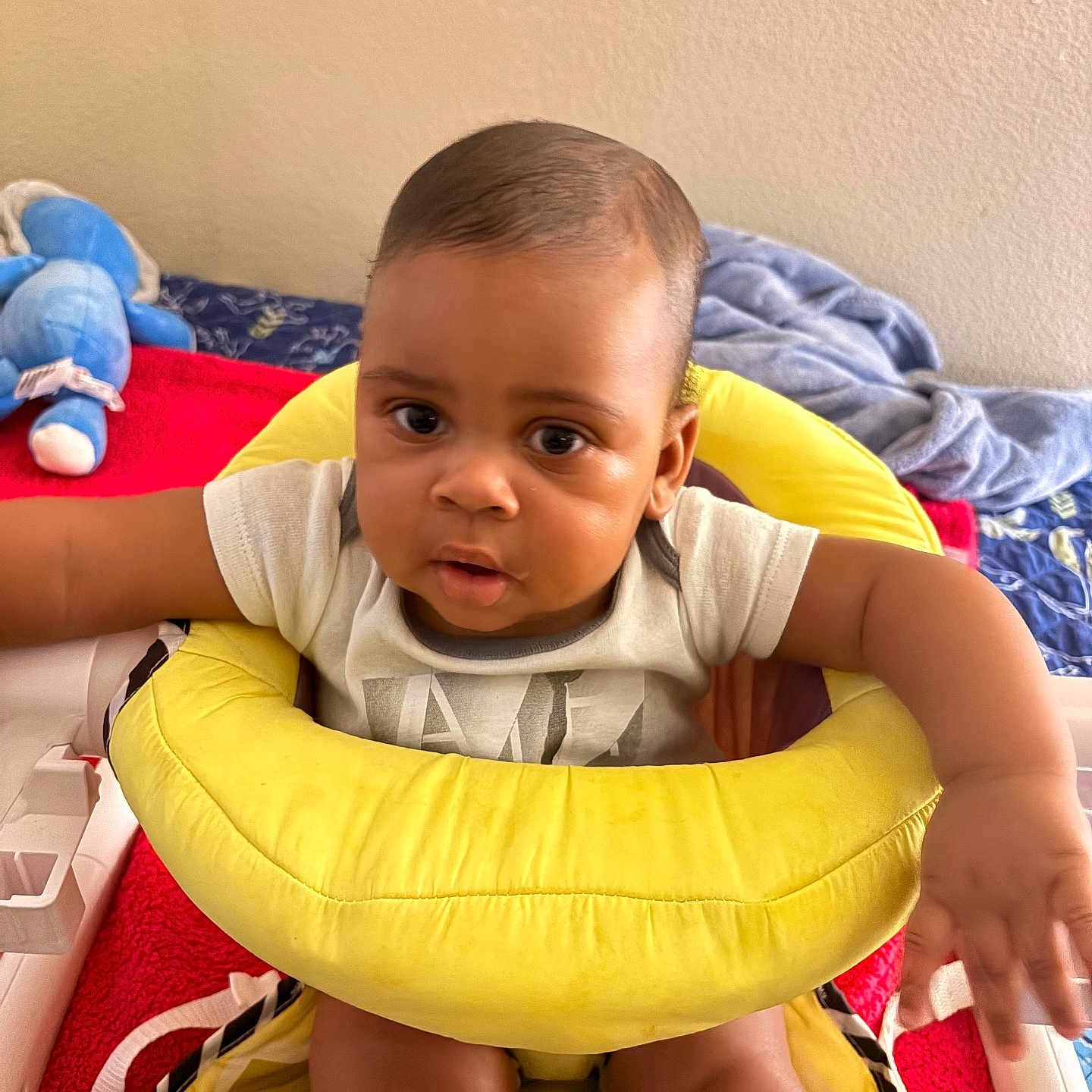 Ean is registered to the contest to win money with this photo: baby, child, infant, yellow_cushion, seat, arm, face, curious, indoor, bed, blanket, stuffed_toy, blue_toy, clothing, person, skin, head, expression, cute, home