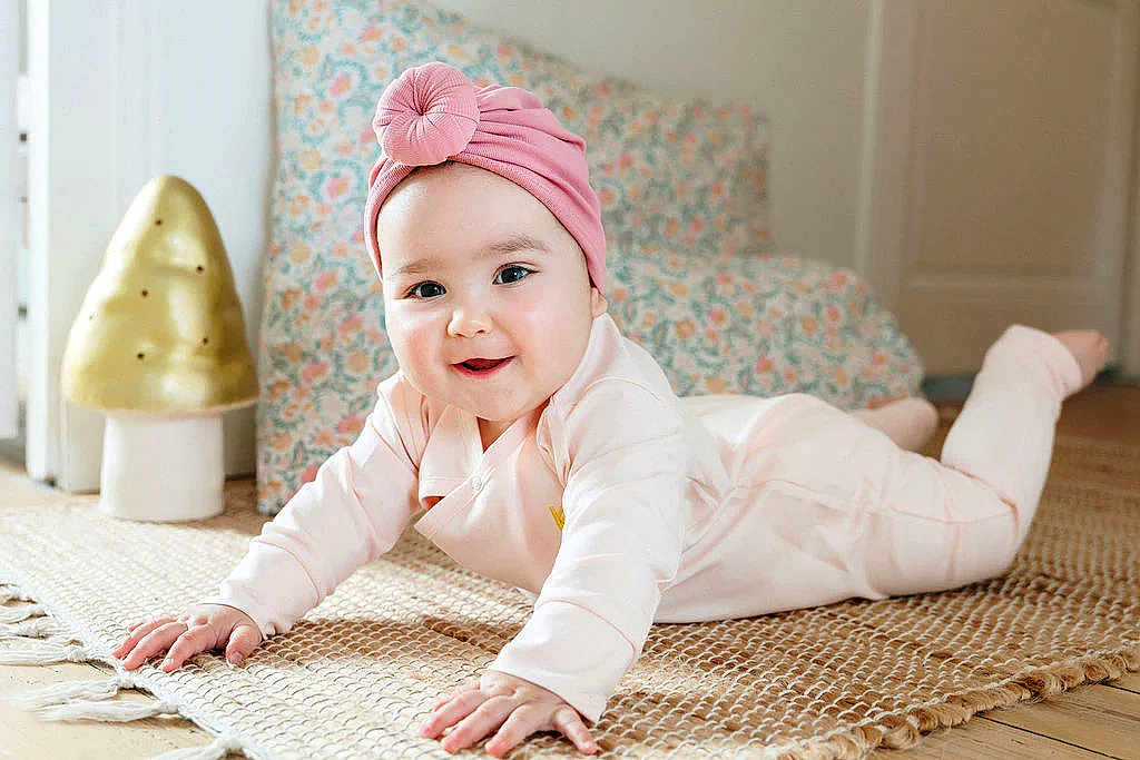 Emy a rejoint le concours — aidez-le/la à gagner de superbes lots ! baby, infant, pink_turban, pink_outfit, smiling, lying_down, hands, rug, woven_rug, floor, decorative_lamp, mushroom_lamp, cushion, floral_pattern, indoor, cozy, child, person, cute, happy