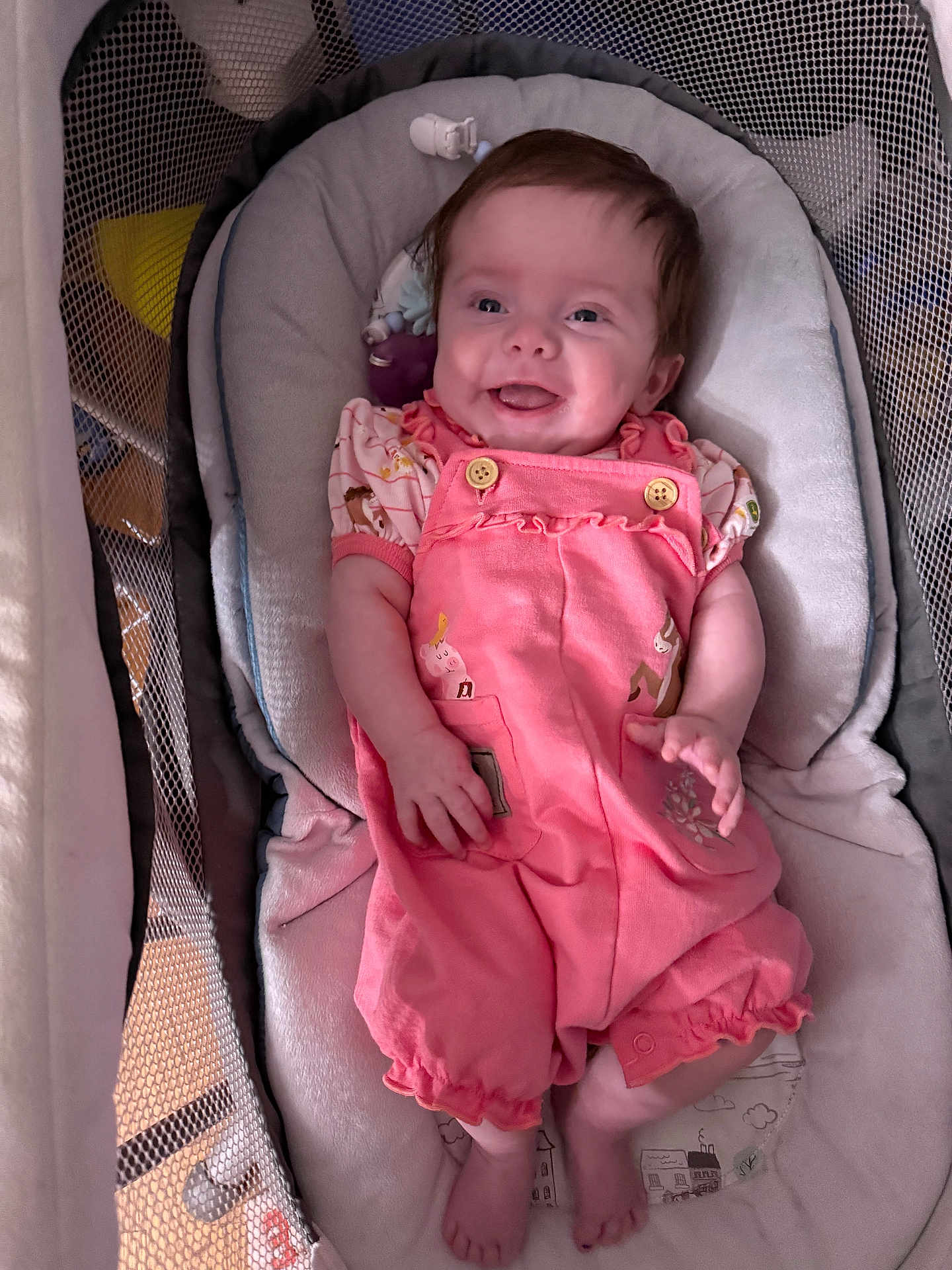 Scarlett is registered to the contest to win money with this photo: baby, infant, smiling, pink_romper, baby_seat, face, person, child, happy, cute, indoor, clothing, legs, feet, padded_seat, mesh_sides, playpen, soft_toys, relaxing, portrait