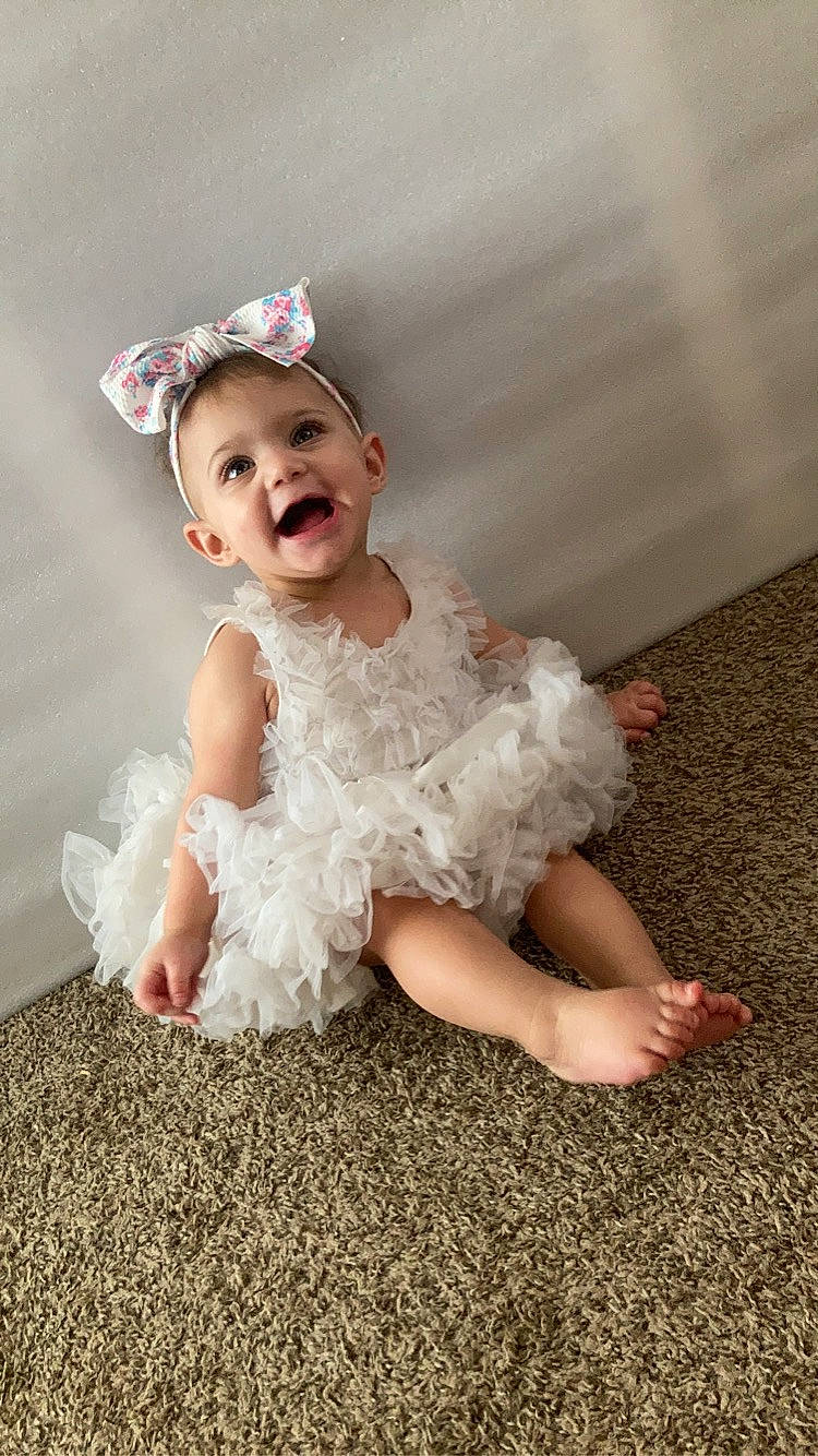 Arielle is registered to the contest to win money with this photo: baby, baby_toddler_clothing, bridal_accessory, child, dress, embellishment, event, fashion_accessory, flash_photography, flooring, fun, happy, headband, headpiece, jewellery, person, pink, ruffle, sitting, smile