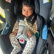 Haven is registered to the contest to win money with this photo: baby, car_seat, child, infant, curly_hair, pacifier, clothing, straps, black, white_outfit, blue_flowers, hands, face, indoors, seat, safety_harness, wheel, floor, expression, person