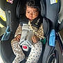 baby, car_seat, child, infant, curly_hair, pacifier, clothing, straps, black, white_outfit, blue_flowers, hands, face, indoors, seat, safety_harness, wheel, floor, expression, person