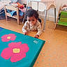 baby, blanket, child, clothing, crawling, curly_hair, floor_mat, furniture, high_chair, indoor, mat, orange_floor, pacifier, patterned_floor, pink_flower, playroom, teal_mat, toddler, toy_bed, wooden_frame
