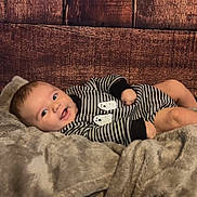 Kolt is registered to the contest to win money with this photo: baby, infant, blanket, striped_clothing, wooden_background, smiling, cozy, cute, face, child, portrait, resting, soft_texture, onesie, happy, young, lying_down, indoor, warm, comfortable