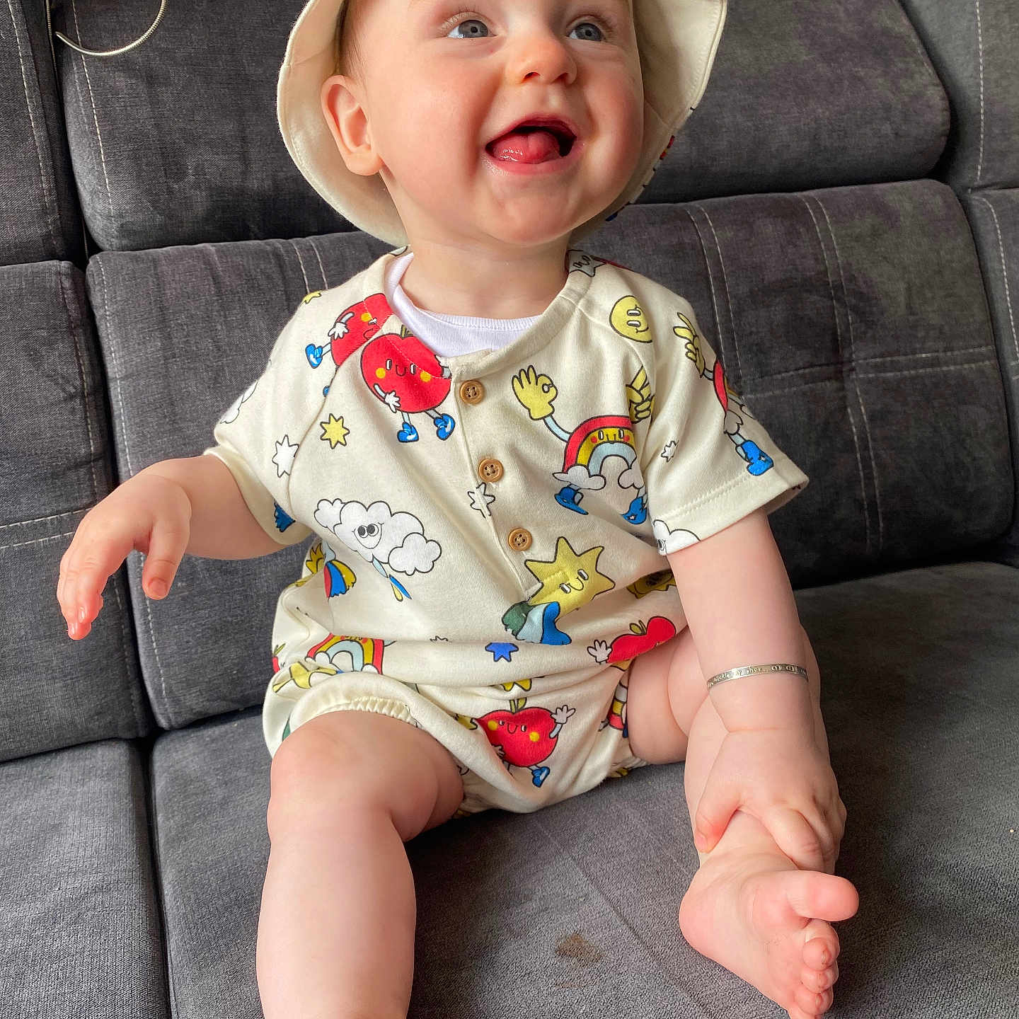 Remi joined the competition — help win amazing prizes! baby, bracelet, child, clothing, couch, cute, face, foot, gray_couch, hand, happy, hat, indoors, infant, person, playful, romper, sitting, smiling, tongue_out