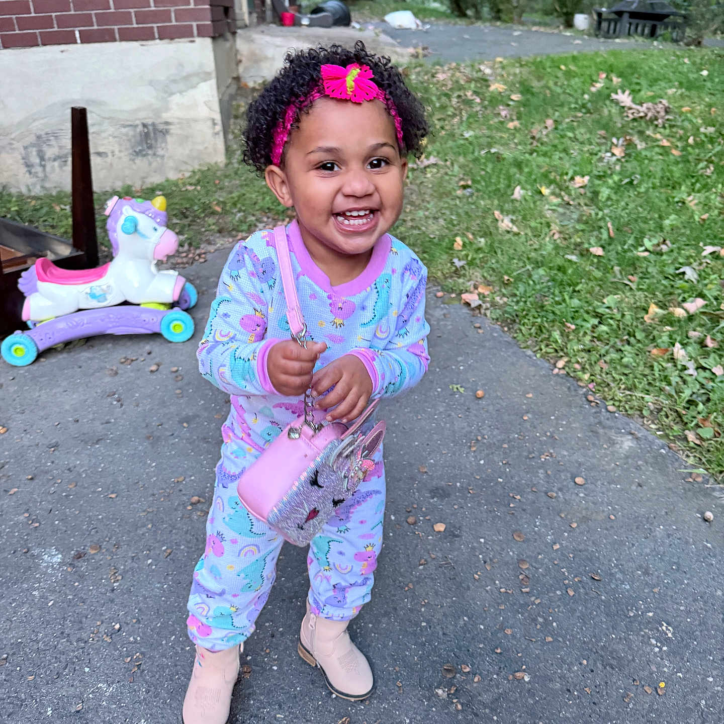 Aimee is registered to the contest to win money with this photo: child, toddler, smiling, happy, outdoors, portrait, curly_hair, headband, pink_clothing, pajamas, boots, purse, toy, unicorn_toy, brick_house, yard, grass, driveway, autumn_leaves, standing