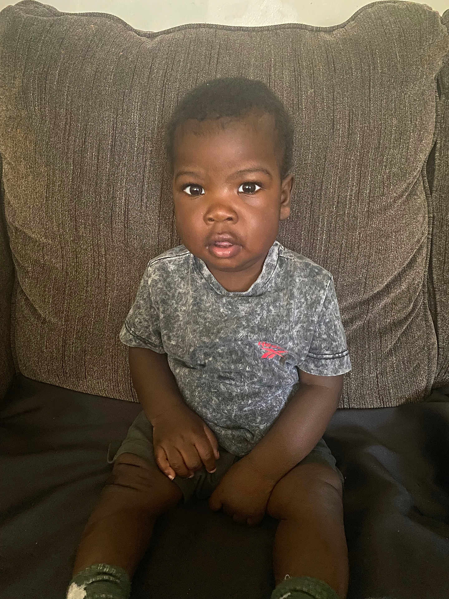 Darren joined the competition — help win amazing prizes! toddler, child, sitting, couch, brown_couch, grey_tshirt, shorts, socks, indoor, portrait, expression, curious, hands, legs, person, young_child, casual_clothing, home, furniture, face