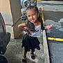 child, girl, smiling, posing, parking_garage, car, concrete, sidewalk, black_skirt, white_top, bow, sandals, long_hair, jewelry, outdoor, happy, standing, person, accessory, parking_lot