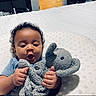 baby, infant, plush_toy, elephant_toy, blue_clothing, cushion, bed, indoor, child, person, smiling, happy, curly_hair, cute, soft_toy, lying_down, comfort, cozy, home, portrait