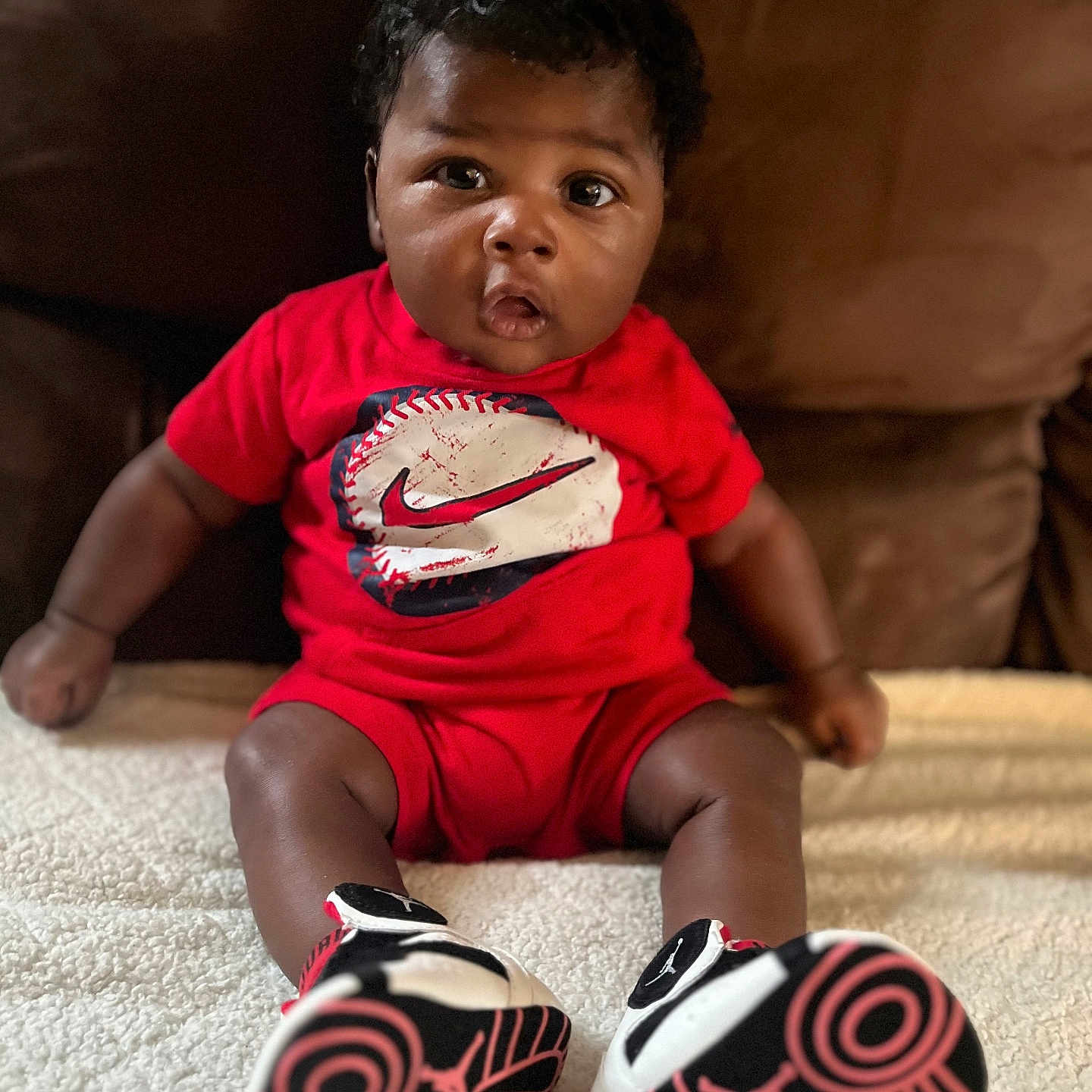 Ashtyn is registered to the contest to win money with this photo: baby, blanket, brown_skin, casual, child, couch, curly_hair, cute, expression, fashion, footwear, indoors, infant, person, portrait, red_clothing, shoes, sitting, sneakers, young_child