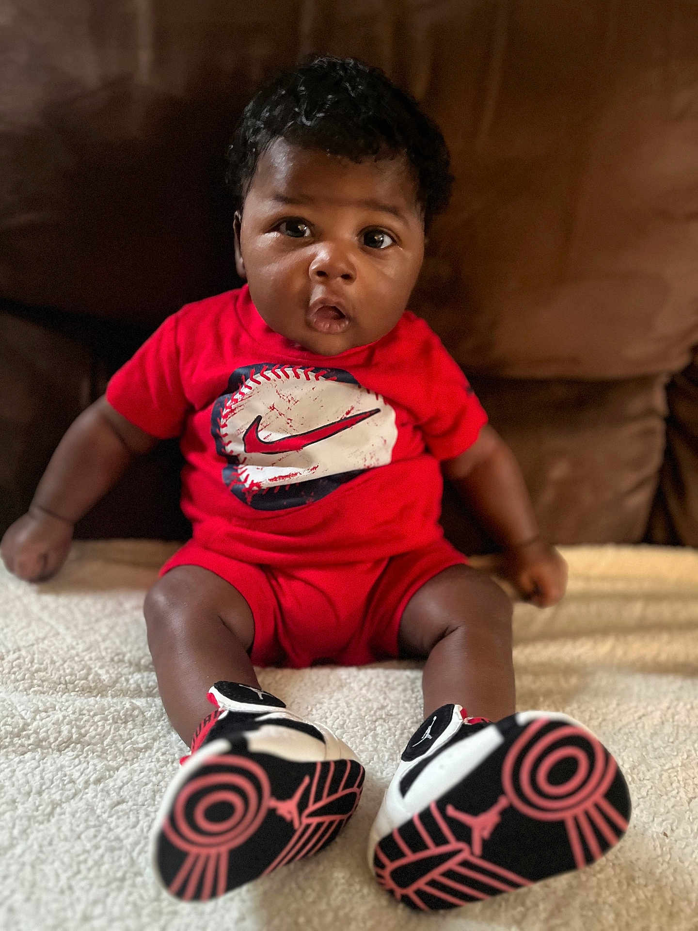 Ashtyn is registered to the contest to win money with this photo: baby, infant, child, red_clothing, sneakers, shoes, sitting, indoors, curly_hair, brown_skin, blanket, couch, portrait, cute, expression, fashion, footwear, young_child, person, casual