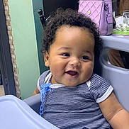 Aïlan a rejoint le concours — aidez-le/la à gagner de superbes lots ! baby, child, high_chair, smiling, curly_hair, striped_clothing, pacifier_clip, indoor, furniture, table, purse, person, cute, happy, face, seated, chubby_cheeks, expression, casual, family