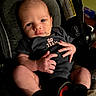 baby, infant, child, car_seat, portrait, face, wide_eyed, sitting, clothing, onesie, team_shirt, adult_hand, hands, legs, socks, indoors, vehicle, safety_label, closeup, photography