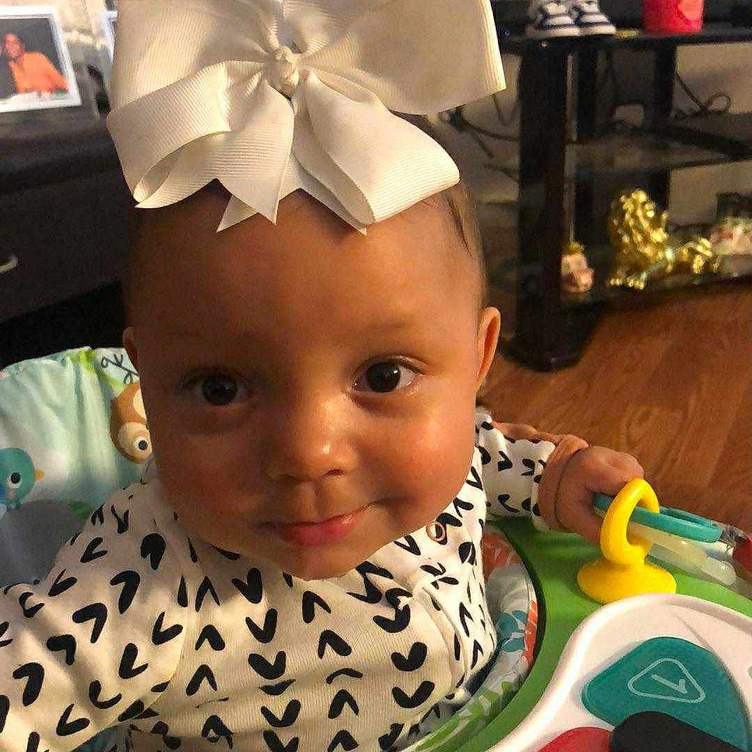 Jordan is registered to the contest to win money with this photo: baby, bow, child, clothing, colorful, cute, eyes, face, floor, furniture, hand, indoor, infant, living_room, person, piano, play, smile, toy, wooden_floor