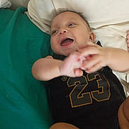 Jordan is registered to the contest to win money with this photo: baby, bed, black_clothing, blanket, child, cute, face, hand_motion, happy, indoors, infant, lying_down, number_23, pillow, portrait, resting, skin, sleepwear, smiling, young