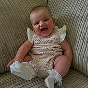 Elsie-Mae joined the competition — help win amazing prizes! baby, infant, smile, laughing, couch, knitted_outfit, socks, frills, happy, cute, indoor, child, person, seated, texture, beige, cozy, portrait, young, comfortable