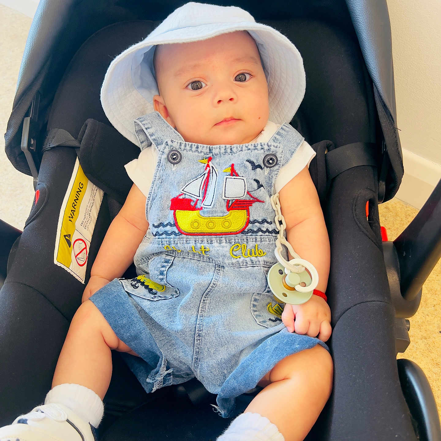 Raym is registered to the contest to win money with this photo: baby, baby_accessory, car_seat, casual, child, clothing, comfortable, cute, denim_overalls, expression, indoor, infant, pacifier, person, portrait, seatbelt, sneakers, socks, white_hat, young_child