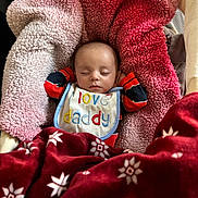 Daniel is registered to the contest to win money with this photo: baby, sleeping, bib, blanket, red_blanket, pink_blanket, cozy, infant, face, closed_eyes, clothing, soft_texture, pattern, indoor, cute, toddler, resting, person, warm, comfort