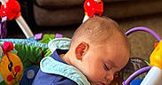 Daniel is registered to the contest to win money with this photo: baby, child, sleeping, activity_center, toy, indoor, colorful, face, head, clothing, blue_shirt, play, furniture, couch, infant, person, rest, peaceful, cute, young