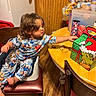 Joseph is registered to the contest to win money with this photo: toddler, child, pajamas, high_chair, table, grinch, gift_box, kitchen, snacks, wood_floor, chair, reaching, curious, indoor, person, clothing, food, container, cabinet, furniture