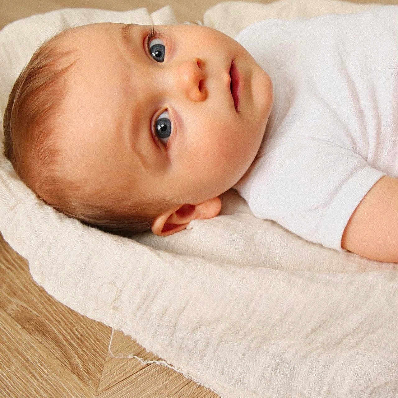 Gaspard participe au concours pour gagner de l'argent avec cette photo : awake, baby, blanket, blue_eyes, cheeks, child, closeup, ear, gaze, head, infant, lying_down, minimalist, mouth, newborn, portrait, skin, soft_texture, white_onesie, wood_floor