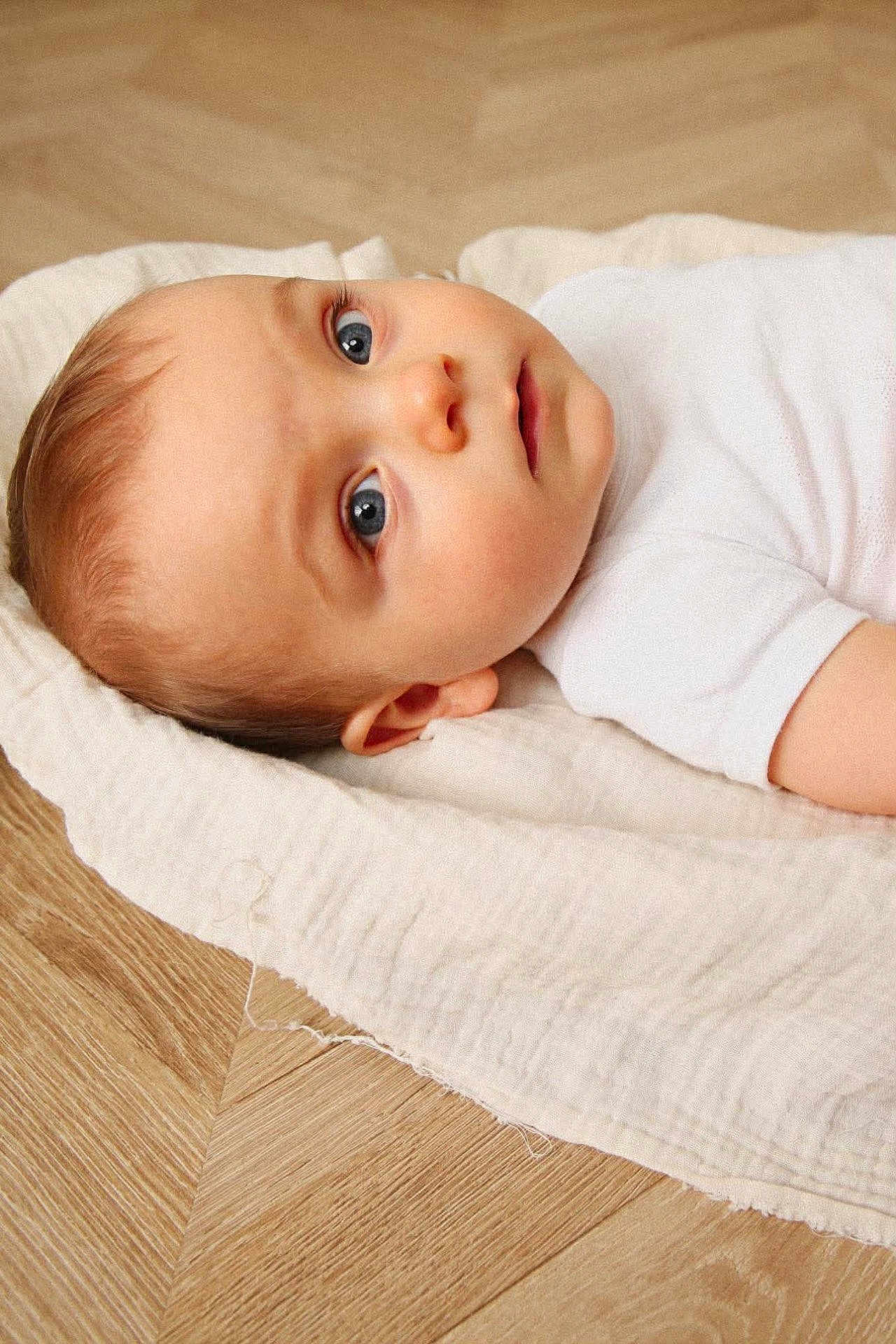 Gaspard participe au concours pour gagner de l'argent avec cette photo : baby, infant, child, blue_eyes, lying_down, white_onesie, blanket, wood_floor, portrait, closeup, cheeks, gaze, ear, skin, soft_texture, awake, mouth, head, newborn, minimalist