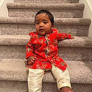 Yeshveer is registered to the contest to win money with this photo: baby, barefoot, carpet, child, close_up, eyes, face, fashion, floral_clothing, hair, home, indoors, infant, photography, portrait, red_outfit, sitting, smiling, staircase, traditional_clothing