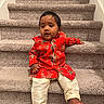 baby, barefoot, carpet, child, close_up, eyes, face, fashion, floral_clothing, hair, home, indoors, infant, photography, portrait, red_outfit, sitting, smiling, staircase, traditional_clothing
