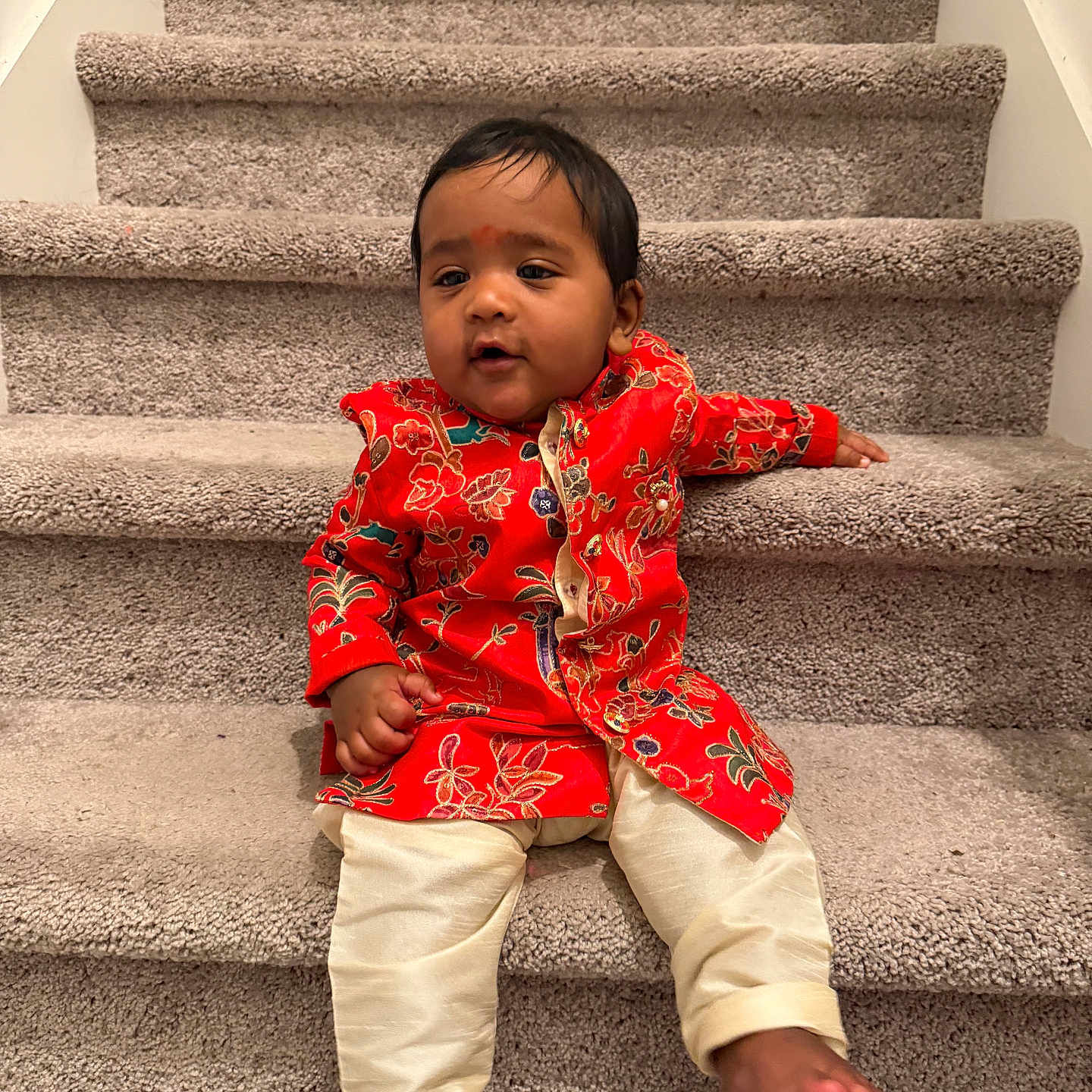 Yeshveer is registered to the contest to win money with this photo: baby, barefoot, carpet, child, close_up, eyes, face, fashion, floral_clothing, hair, home, indoors, infant, photography, portrait, red_outfit, sitting, smiling, staircase, traditional_clothing